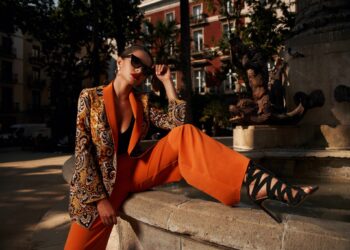 Fashion style outdoor portrait. Elegant woman in colorful orange costume, sunglasses and black shoes sitting on stone fence of fountain on a sunny evening in tropical city