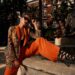 Fashion style outdoor portrait. Elegant woman in colorful orange costume, sunglasses and black shoes sitting on stone fence of fountain on a sunny evening in tropical city