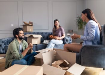 Friends sitting and talking in their new apartment while unpacking their things from cardboard box.