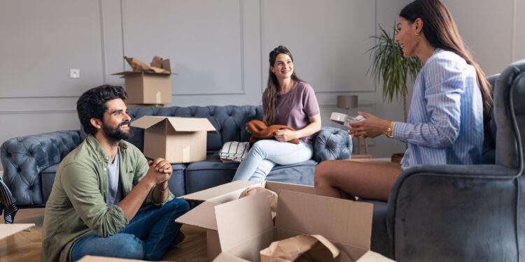 Friends sitting and talking in their new apartment while unpacking their things from cardboard box.