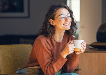 Happy young woman drinking a cup of tea in an autumn morning. Dreaming girl sitting in living room with cup of hot coffee enjoying under blanket with closed eyes. Pretty woman wearing sweater at home and enjoy a ray of sunshine on a winter afternoon.