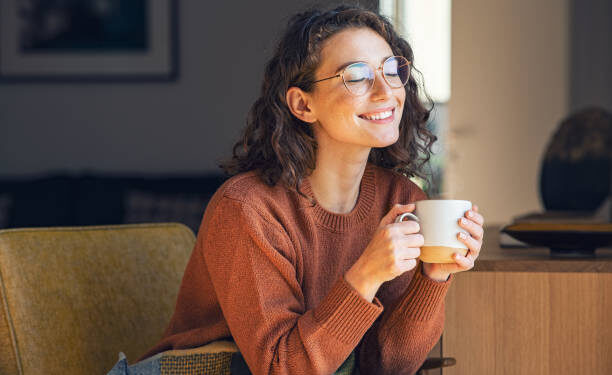 Happy young woman drinking a cup of tea in an autumn morning. Dreaming girl sitting in living room with cup of hot coffee enjoying under blanket with closed eyes. Pretty woman wearing sweater at home and enjoy a ray of sunshine on a winter afternoon.