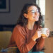 Happy young woman drinking a cup of tea in an autumn morning. Dreaming girl sitting in living room with cup of hot coffee enjoying under blanket with closed eyes. Pretty woman wearing sweater at home and enjoy a ray of sunshine on a winter afternoon.